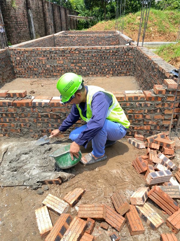 Người thợ đang chuẩn bị dùng bay ải đều vữa lên bề mặt tường và tiếp tục đặt các viên gạch đỏ (đang xếp đống xung quanh) để nâng cao bức tường móng