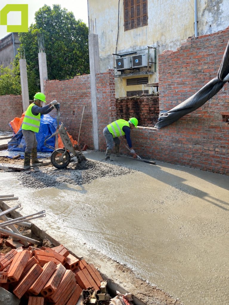 Hai người thợ thi công đang thực hiện công đoạn đổ và làm phẳng bề mặt bê tông nền.