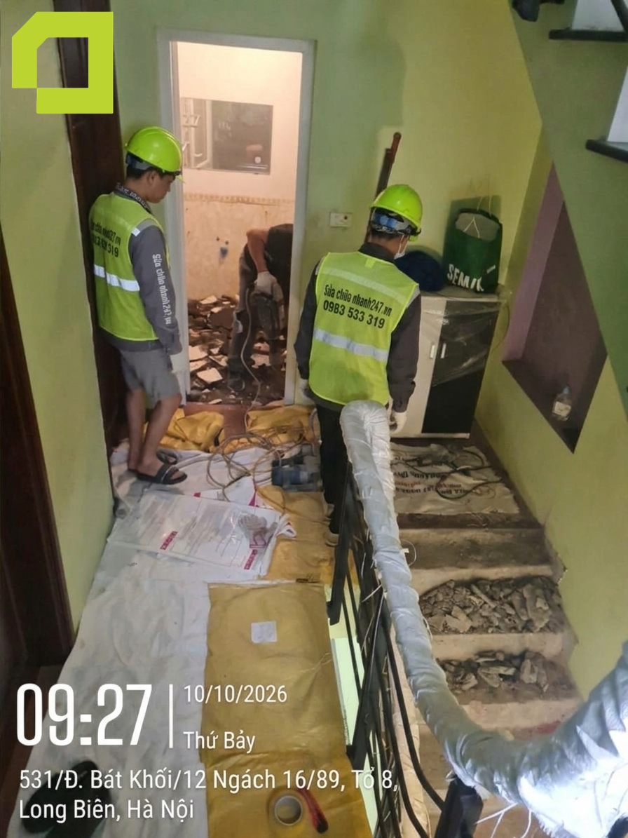 Hai thợ ở ngoài khu vực nhà vệ sinh đang quan sát, hỗ trợ hoặc chuẩn bị vật tư.